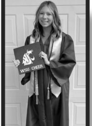 A black and white photo of Mandy, a young woman with fare skin and light curled hair, wearing a graduation gown, sash, and cords. She stands smiling while holding a sign on the WSU logo that ssy WSU Cheer.